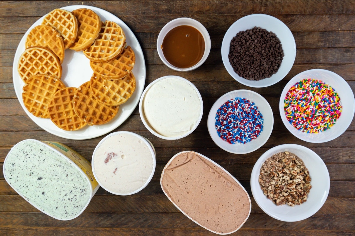 A plate with waffles, a bowl with various ice cream toppings, and different ice cream flavors prepared on a table.