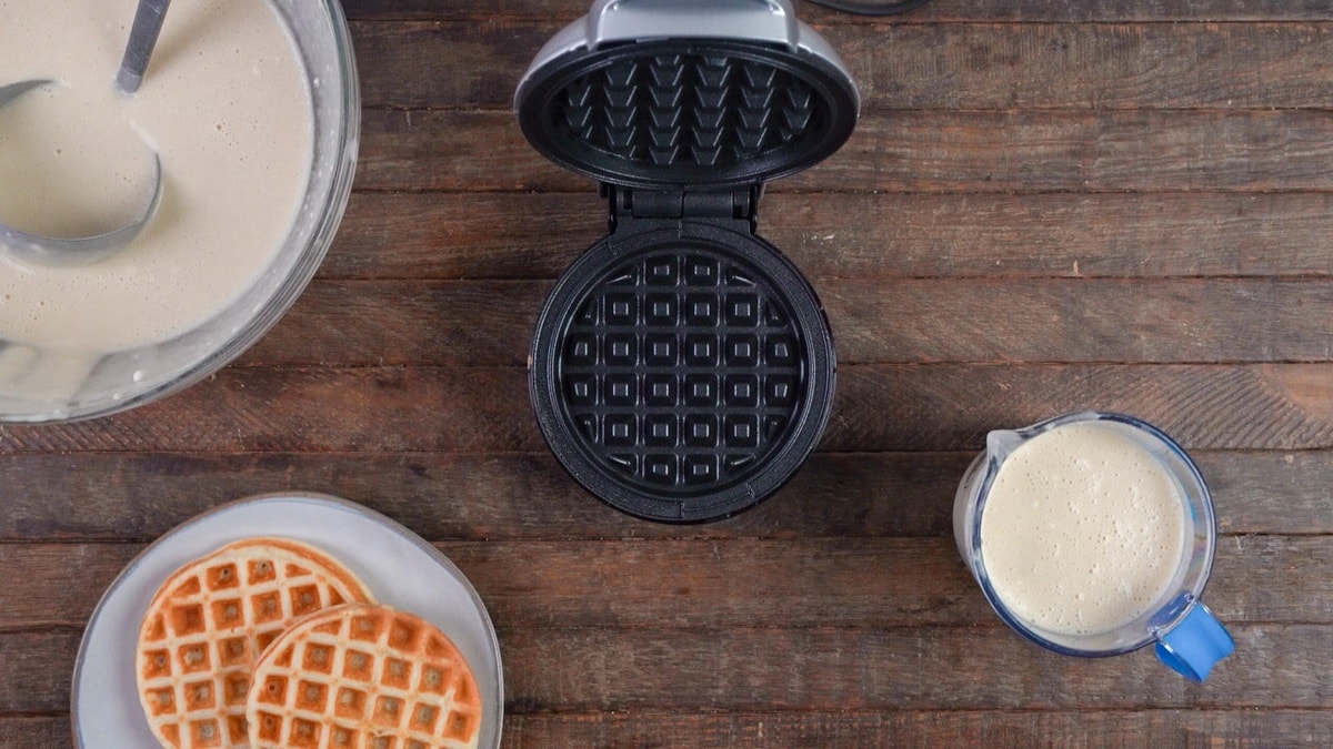 Waffle batter and an open waffle iron, ready for use, on a table.