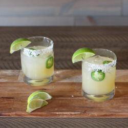 Two glasses of Jalapeño Margarita with lime wedges on a wooden surface.