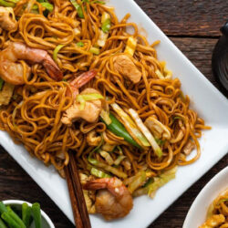 A serving of Mee Goreng with shrimp, chicken, and vegetables on a wooden table.