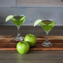 Two glasses of sour apple martini and two apples are placed on a table.